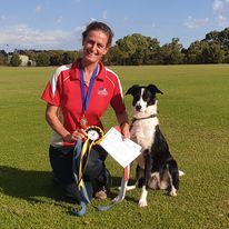 A woman in a red and white shirt kneels beside a black and white dog on a grassy field, holding a medal and a certificate.